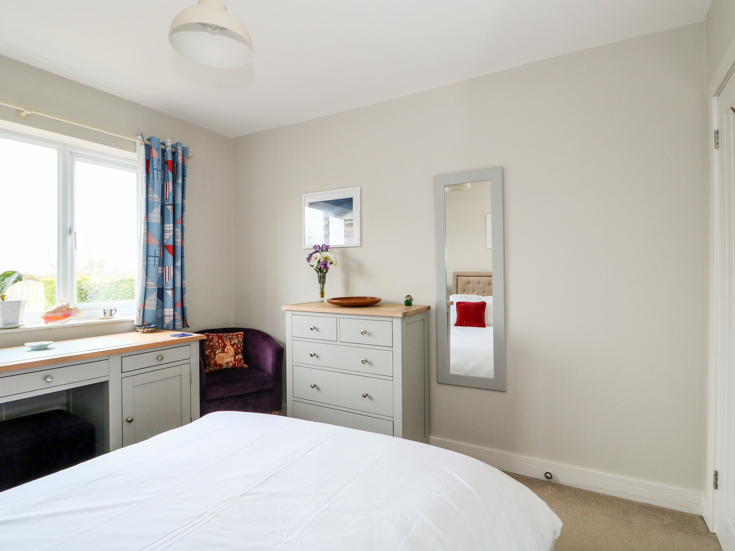 A bedroom with a bed, a dresser, a desk, a chair, a window with curtains, a mirror, and a view of the outdoors.