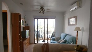 Ein Wohnzimmer mit einer Couch, einem Esstisch und einem Fernseher. Eine Deckenventilator und eine Klimaanlage sind an der Wand angebracht.