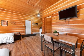 A wooden cabin interior with a bed, a dining table with chairs, a TV mounted on the wall, and a small plant on the table. - Paddlers Inn - Canoe Shop | Rustic | Quaint | Cozy (Canal Flats)