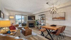 A modern living room with a fireplace, a dining area with a wooden table and chairs, and a large window offering a view of the outdoors. - Mountain Modern | Outdoor Pool | Hot Tub | Gym (Invermere)