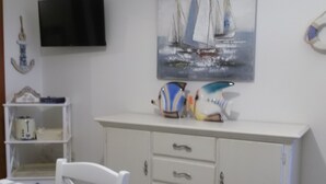 Un coin repas avec une table et des chaises blanches, un meuble blanc avec des tiroirs, un téléviseur à écran plat fixé au mur et un tableau représentant des voiliers, placé au-dessus du meuble.