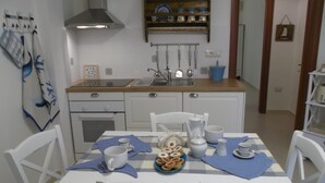 Une petite cuisine avec des meubles blancs, une cuisinière et un évier. On y trouve une table avec des chaises, un bol rempli de beignets et une machine à café.