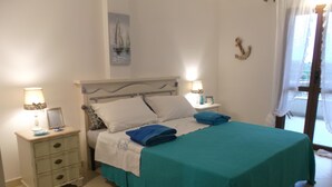 Un lit bien fait, recouvert d’une courtepointe turquoise, avec deux oreillers et une serviette bleue pliée. Il y a deux tables de chevet avec des lampes et un petit tableau encadré. Un tableau représentant des voiliers est accroché au mur au-dessus du lit.