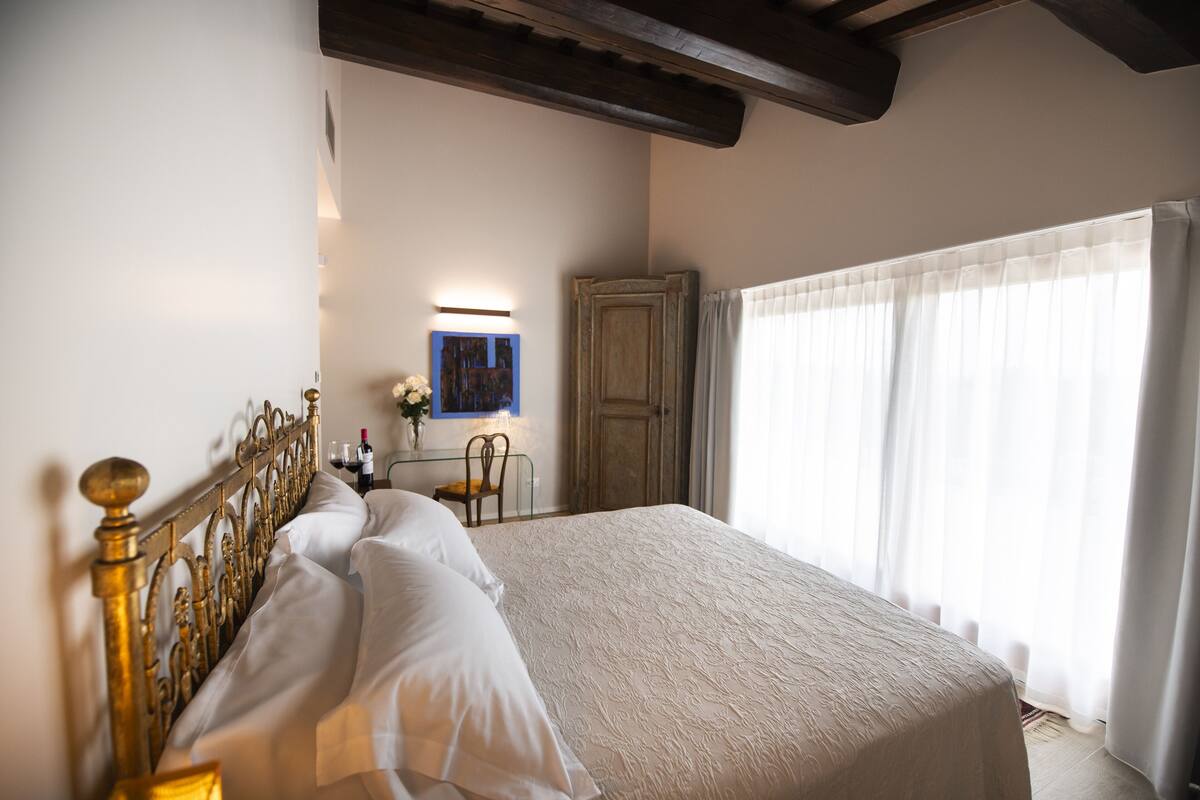 A neatly made bed with a headboard, a bedside table with a vase of flowers, and a window with sheer curtains.