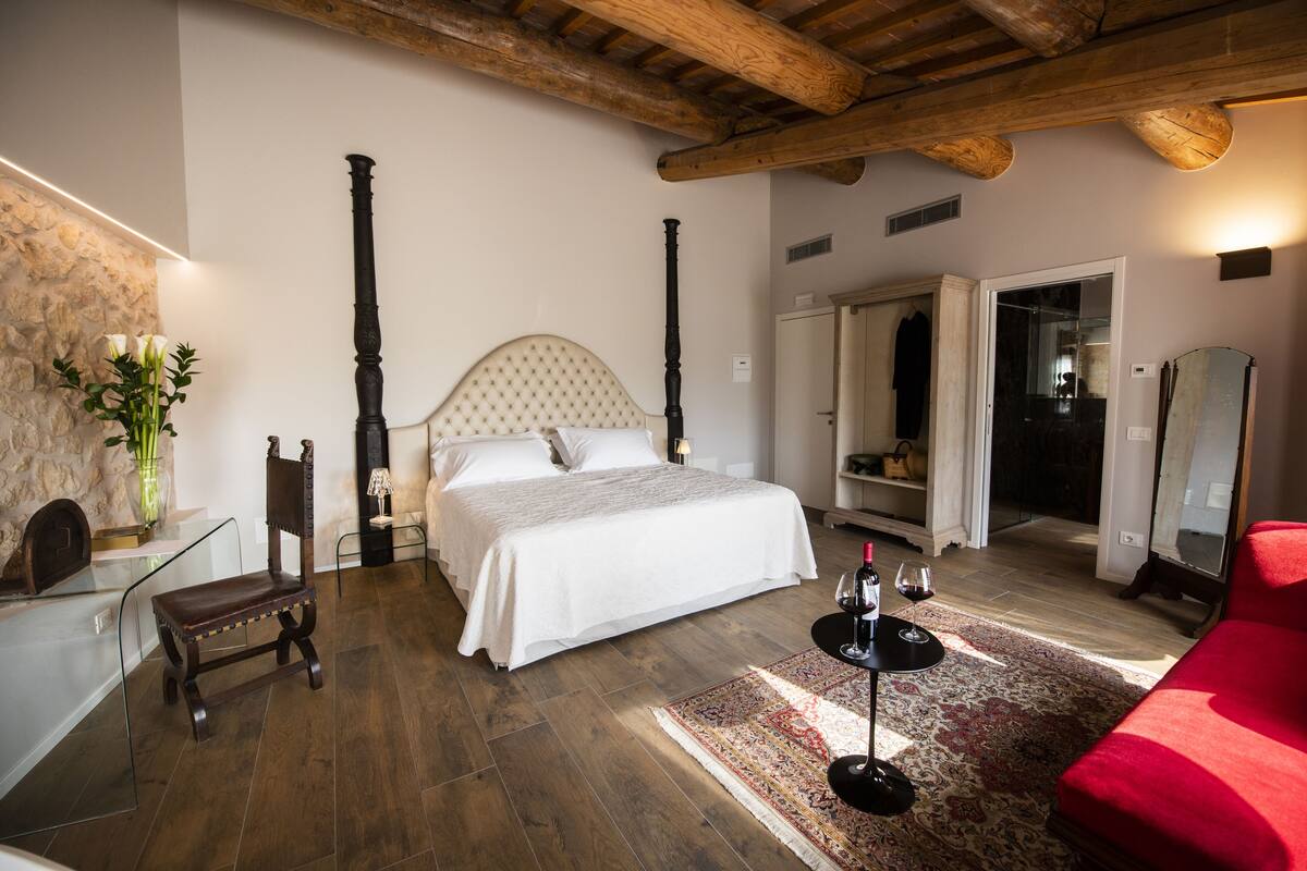 A bedroom with a large bed, a wooden chair, a red sofa, a glass table with a wine glass, and a mirror.