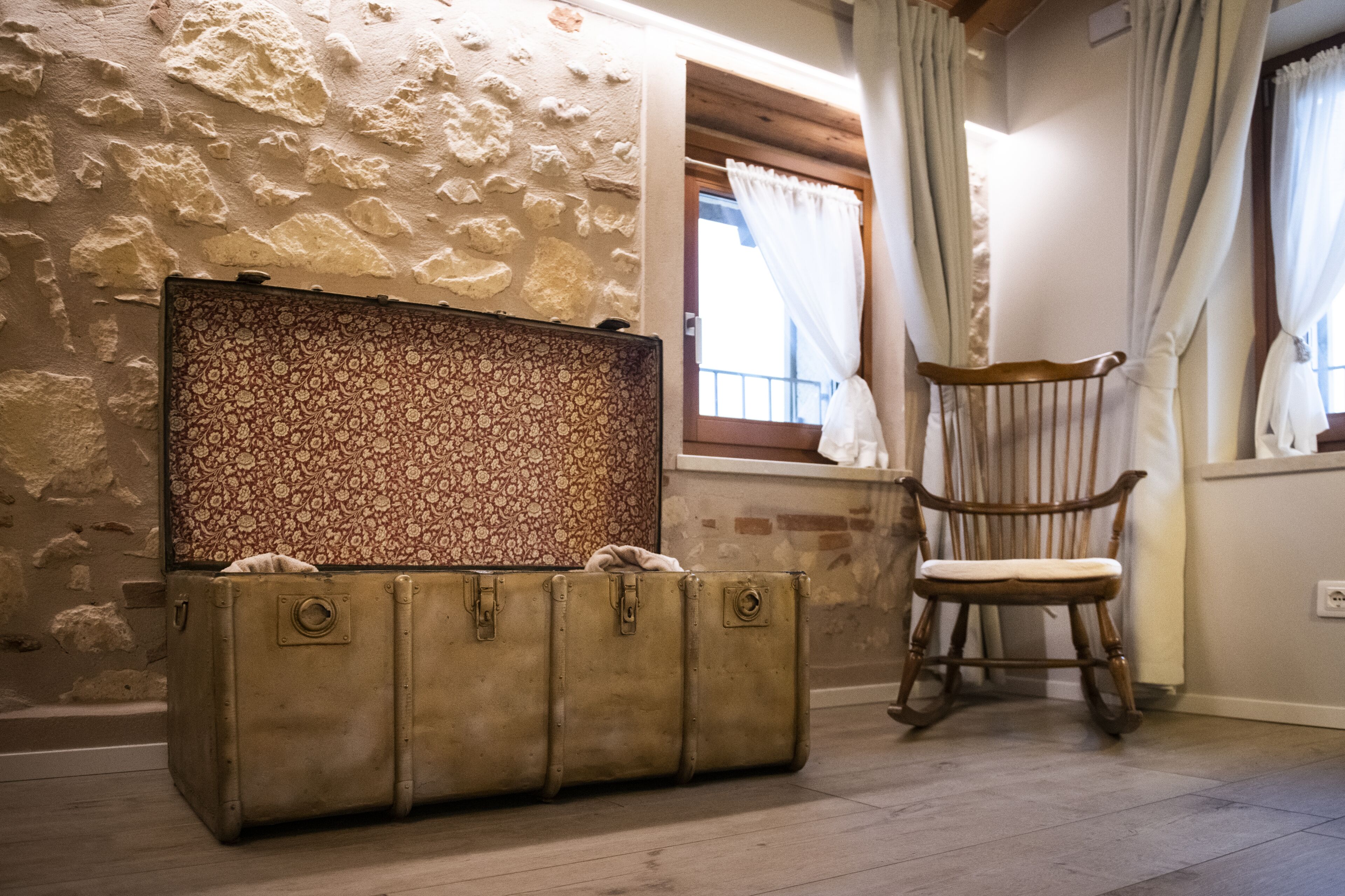 A room with a vintage suitcase, a wooden rocking chair, and a stone wall.