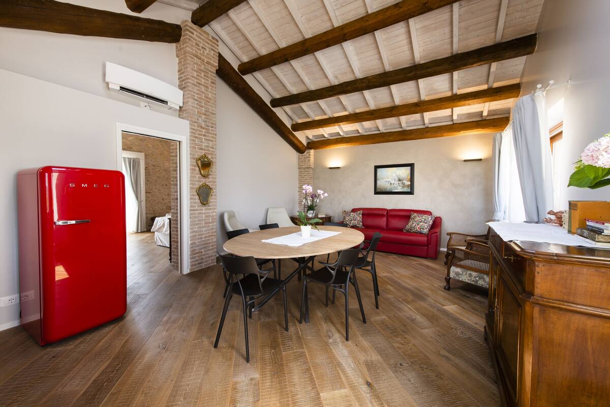 A dining area with a wooden table, chairs, a red refrigerator, a red sofa, and a wooden desk with a plant.