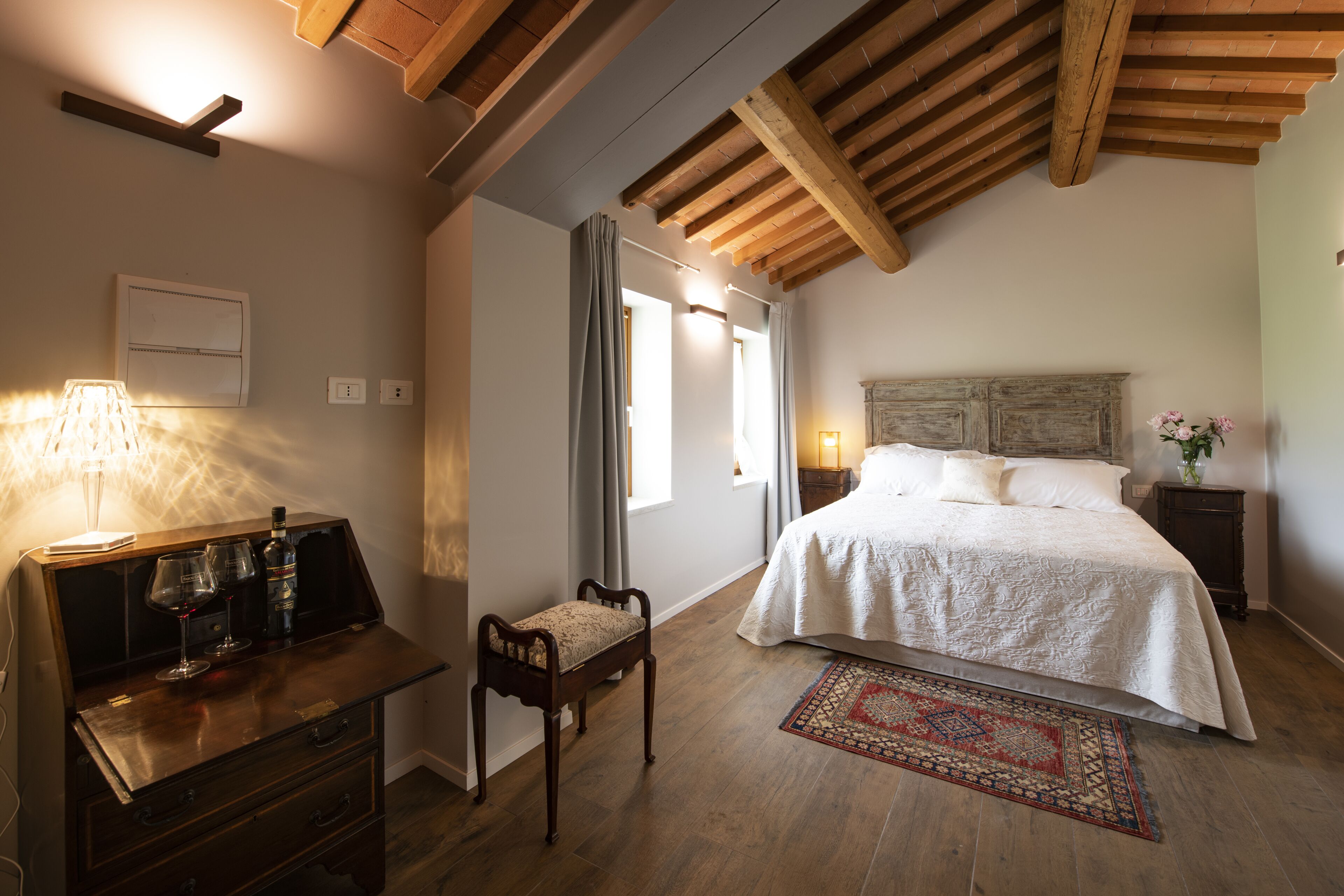 A bedroom with a wooden ceiling, a bed with white bedding, a nightstand with a lamp, and a small bench.