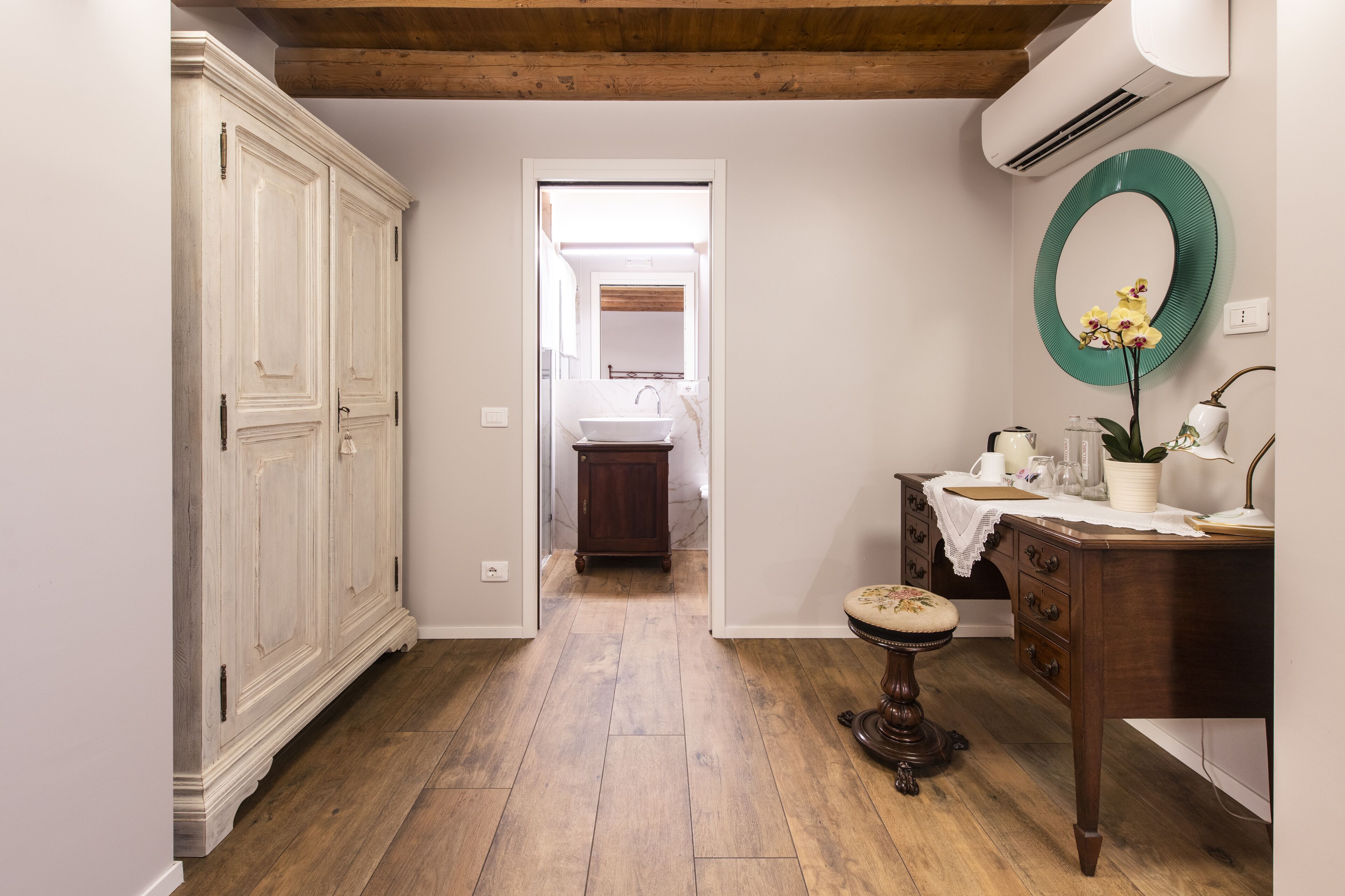 A narrow hallway with a wooden floor, a white wardrobe, a vanity with a mirror, and a small table with a stool.
