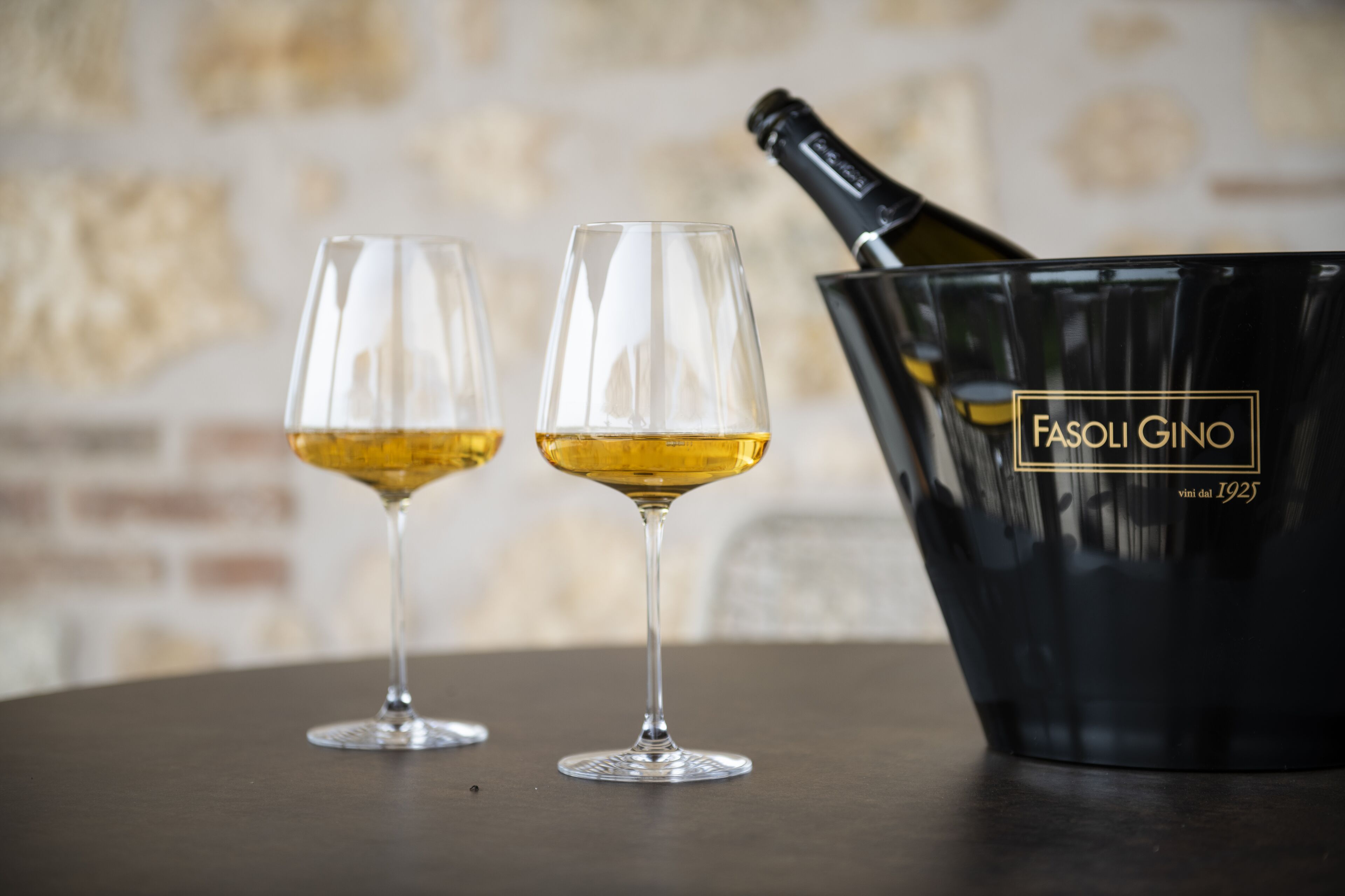 Deux verres de vin blanc et un seau noir portant l’inscription « FASOLI GINO » sont posés sur une table.