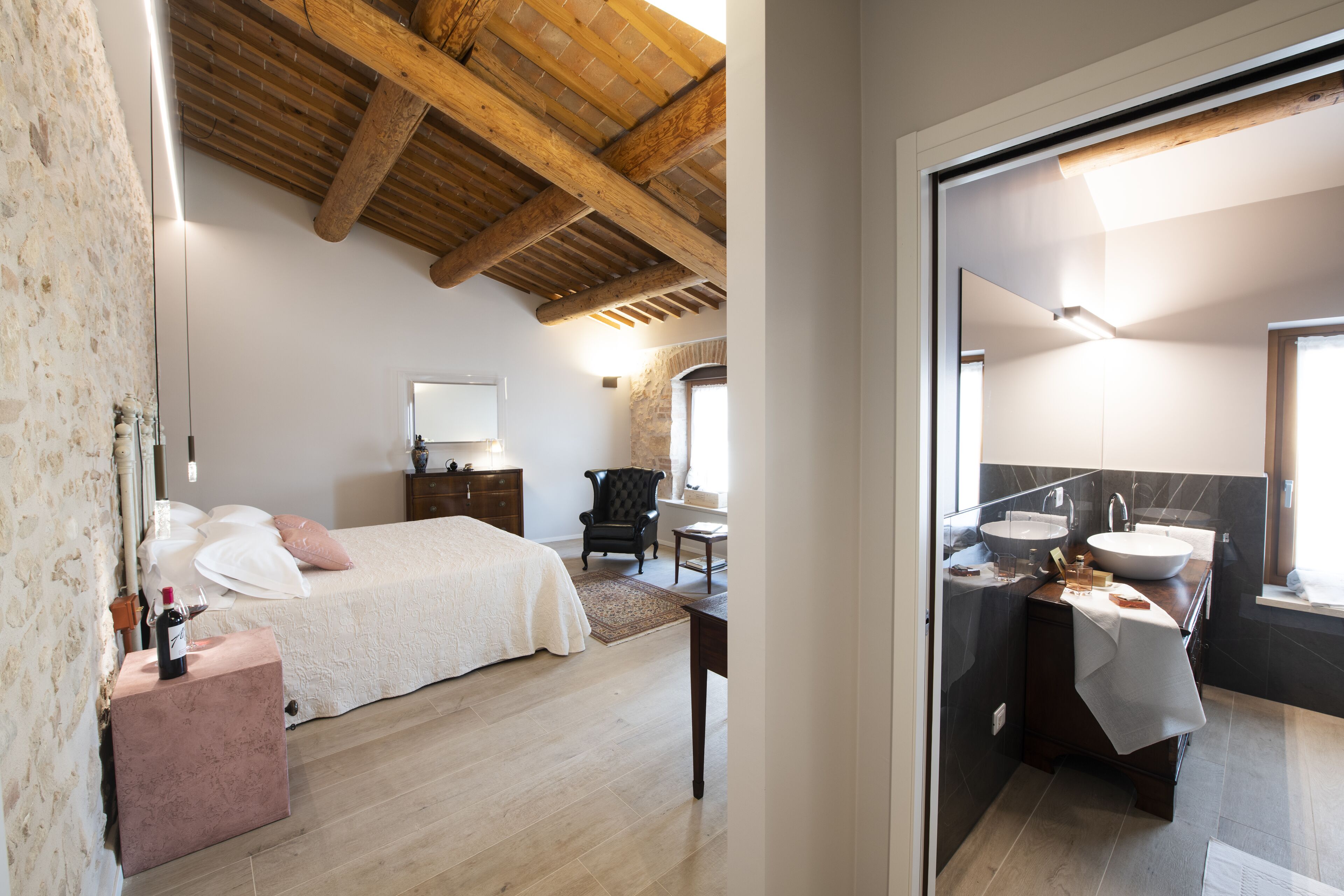 Een slaapkamer met een bed, nachtkastje en stoel. Er is een badkamer met een wastafel en spiegel.