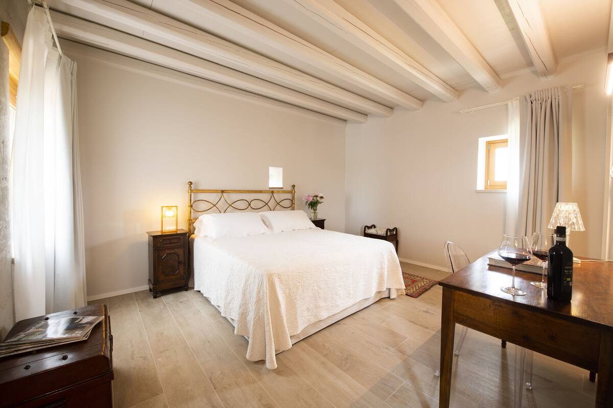 A bedroom with a bed, a nightstand, a dresser, and a table with a lamp and wine glasses.