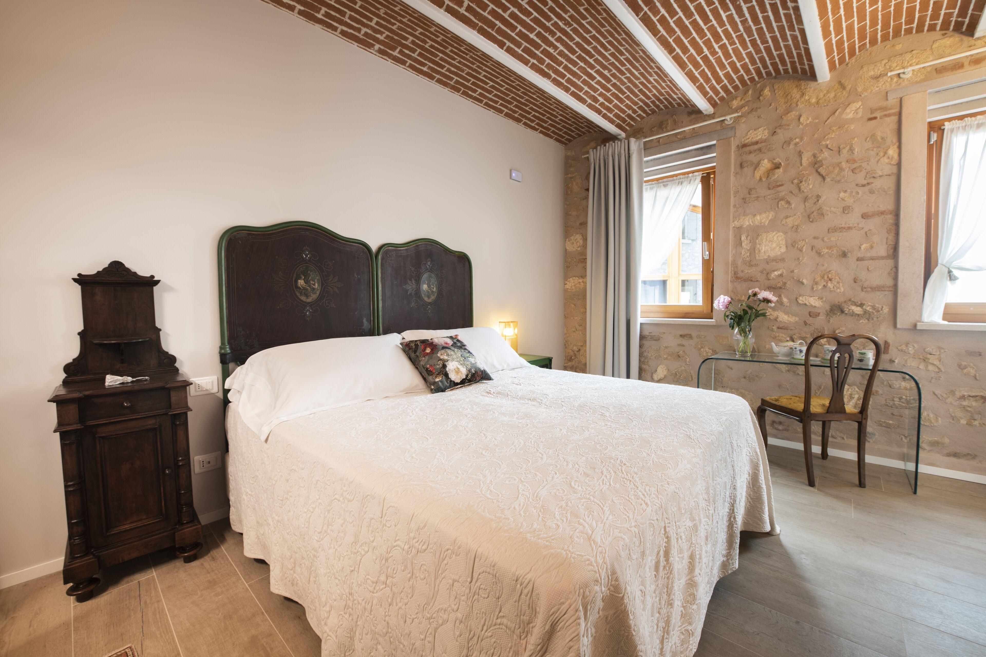 Une chambre avec un grand lit, une table de chevet en bois, une table en verre avec une chaise, et un mur en pierre.