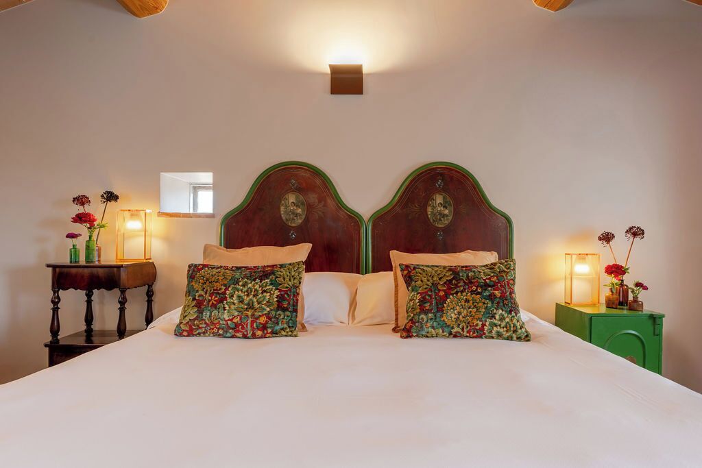 A double bed with floral-patterned pillows, a nightstand with a lamp, and a small table with flowers.