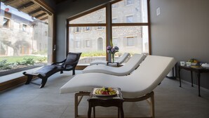 Una habitación iluminada por el sol con dos sillones reclinables, una mesita con un frutero y vistas a un patio a través de amplias ventanas.