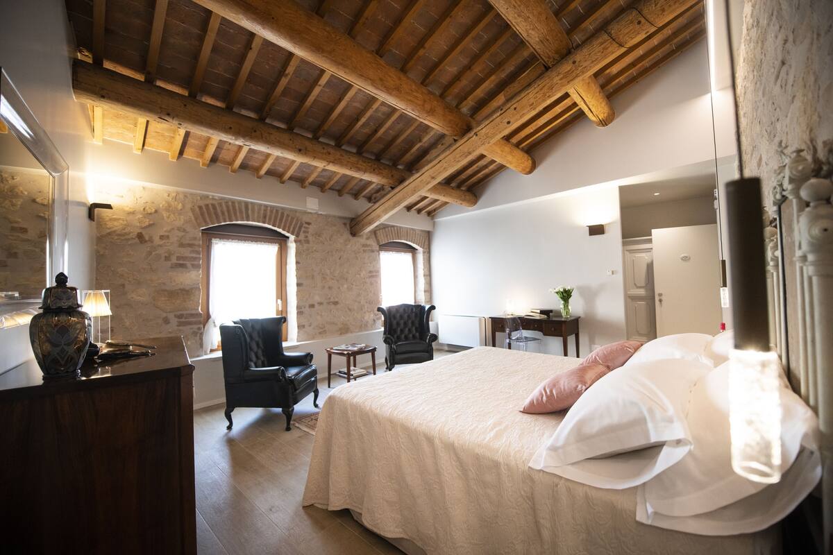 A bedroom with a large bed, wooden ceiling beams, stone walls, a desk, and a chair.