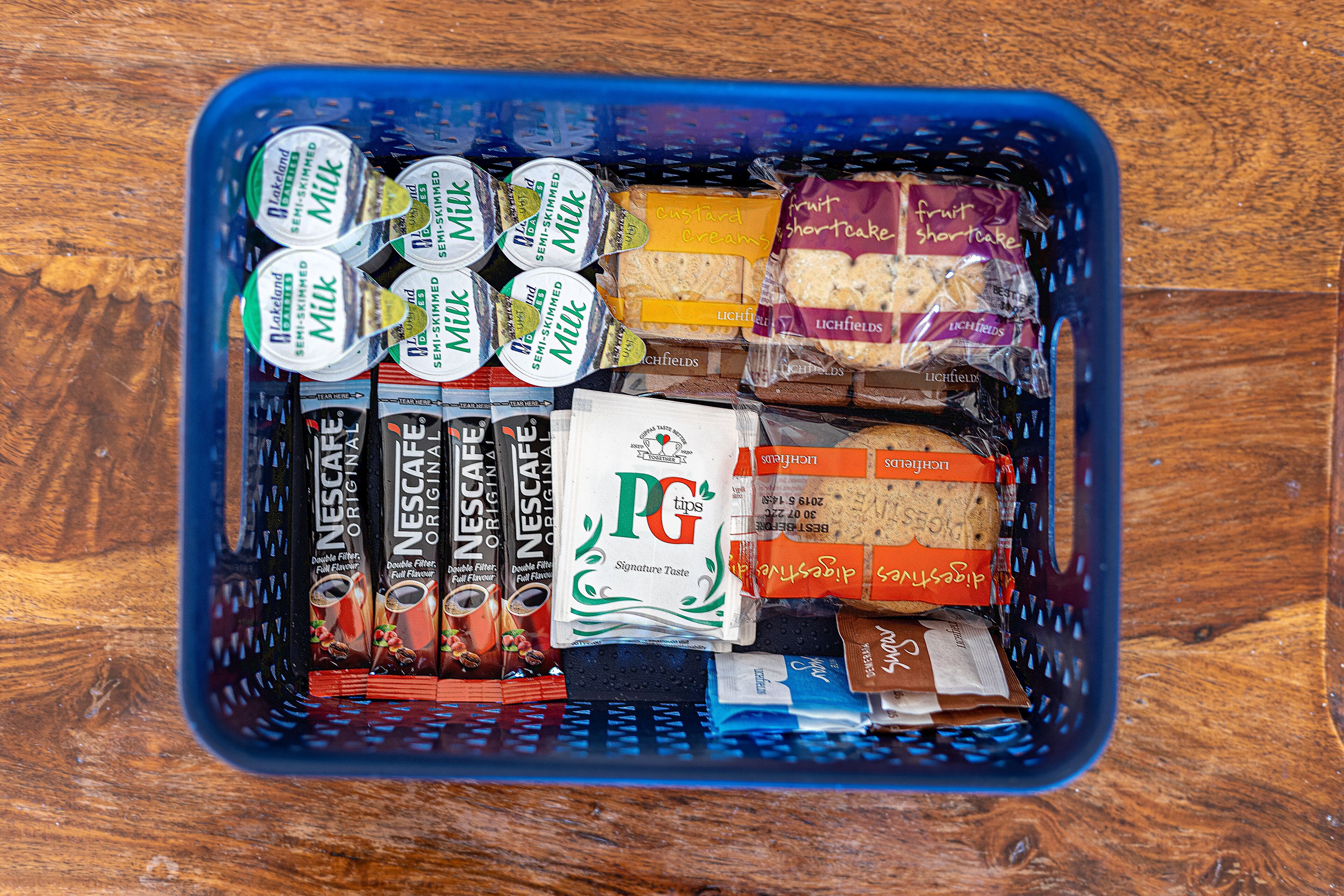 A blue basket filled with various food items including Nescafé Original coffee, PG tips coffee, and different types of cookies and snacks.