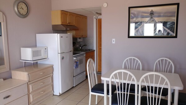 A compact kitchen with a refrigerator, microwave, and wooden cabinets. There is a dining table with four chairs and a framed picture on the wall. - Oceanfront Condo (Wildwood Crest)