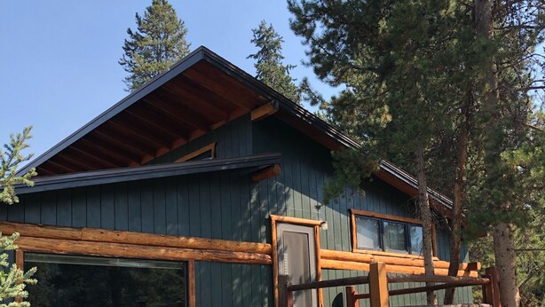 Une cabane en rondins avec une terrasse en bois, un escalier menant à un porche et une grande fenêtre donnant sur une zone boisée.
