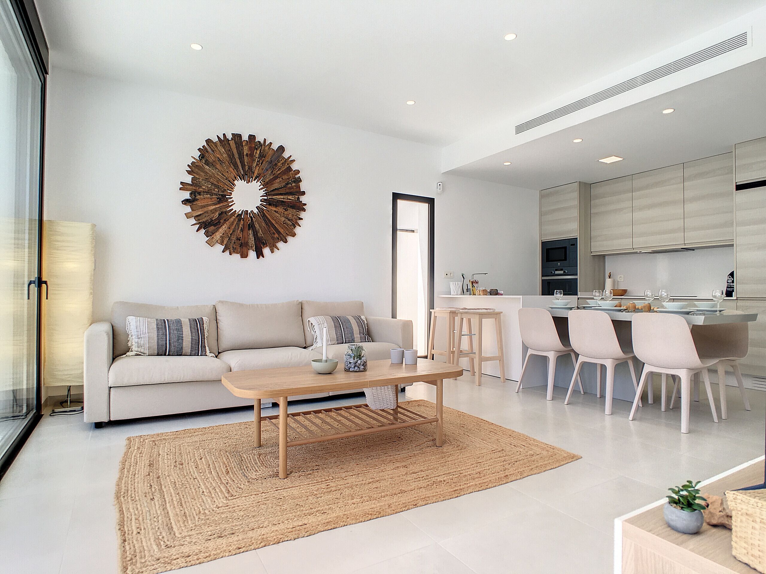 Un salon moderne avec un canapé, une table basse et des fauteuils, une cuisine avec un coin repas et un élément décoratif mural.
