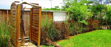 A wooden outdoor shower with a wooden door, surrounded by a garden with various plants and a grassy area.