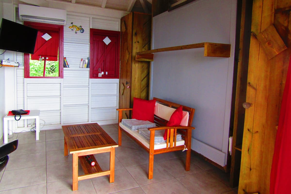 Una habitación con una mesa de madera, un sofá con cojines rojos, un televisor en la pared y una ventana con contraventanas.