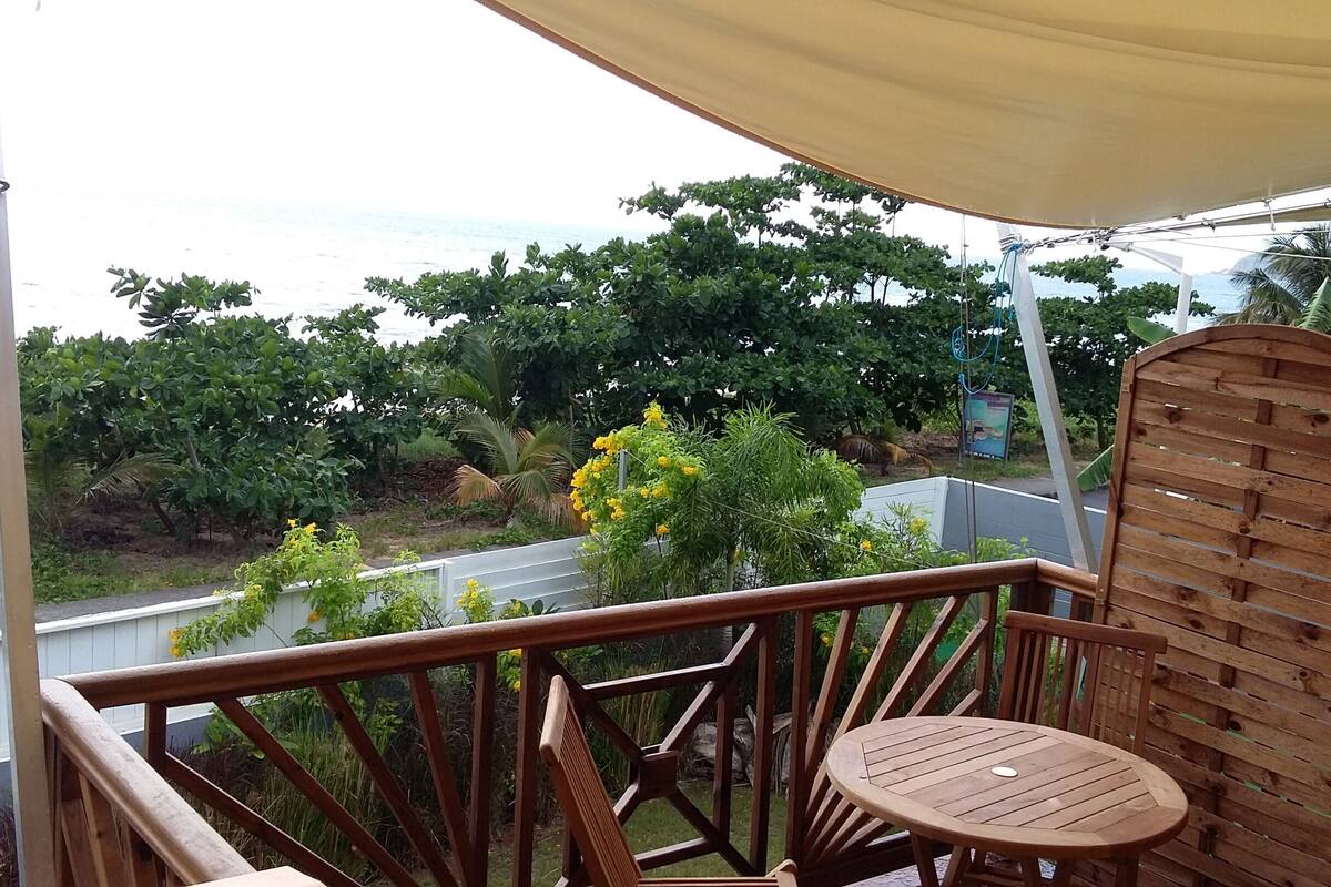 Una terraza de madera con una mesa redonda y una silla de madera, con vistas a un jardín con varios árboles y el mar.