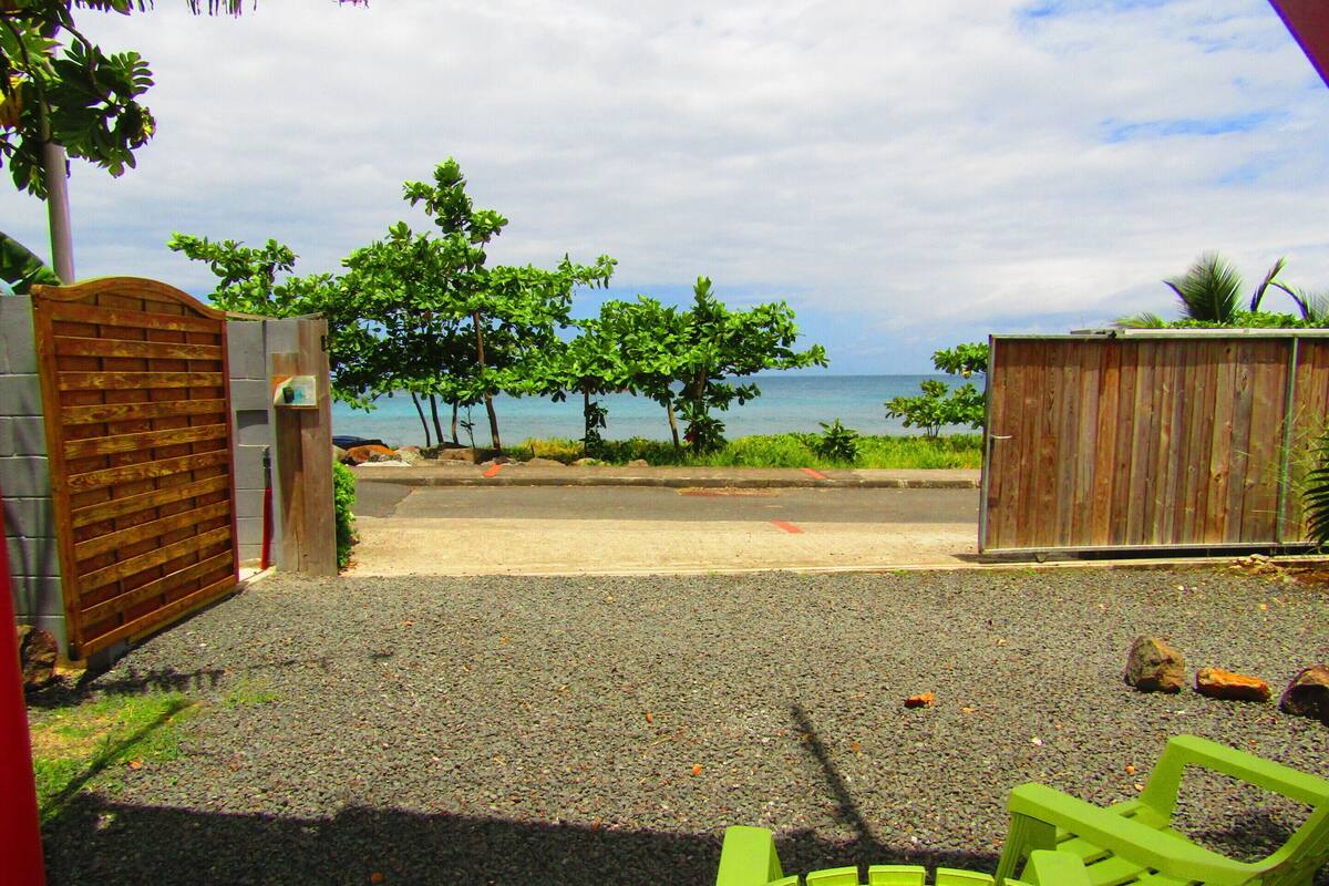 Vista de una playa con una valla de madera, un camino de grava y sillas verdes al frente.