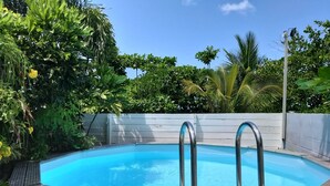 Terraza de madera con piscina, rodeada de abundante vegetación y una valla blanca.