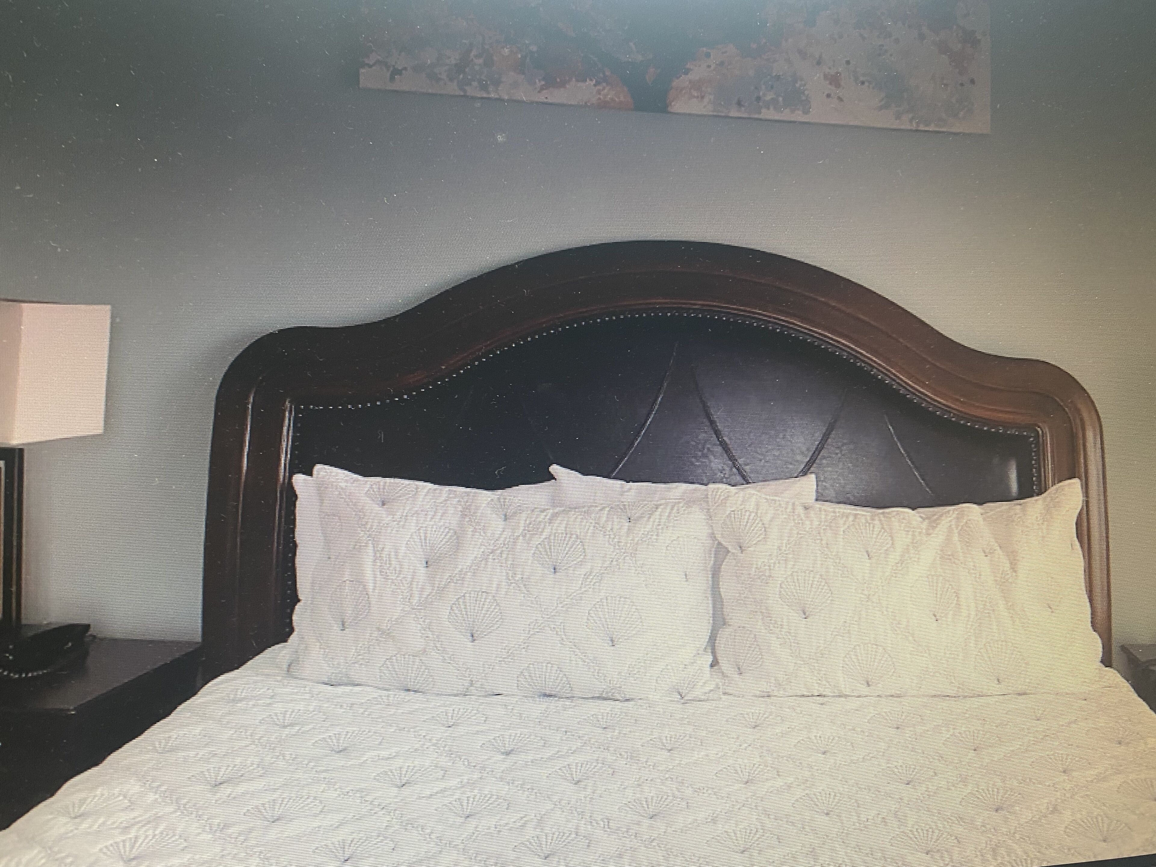 A bed with a dark headboard and white bedding in a room with a plain wall and a painting above the bed.