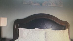 Ein Bett mit dunklem Kopfteil und weißer Bettwäsche in einem Raum mit schlichter Wand und einem Gemälde über dem Bett.