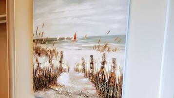 A framed painting of a beach scene with a path leading to the sea, featuring reeds and a sailboat in the distance.