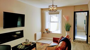 A living room with a TV, a fireplace, a window with blinds, a sofa, and a plant. - Tralee Townhouse Holiday Home (Tralee)
