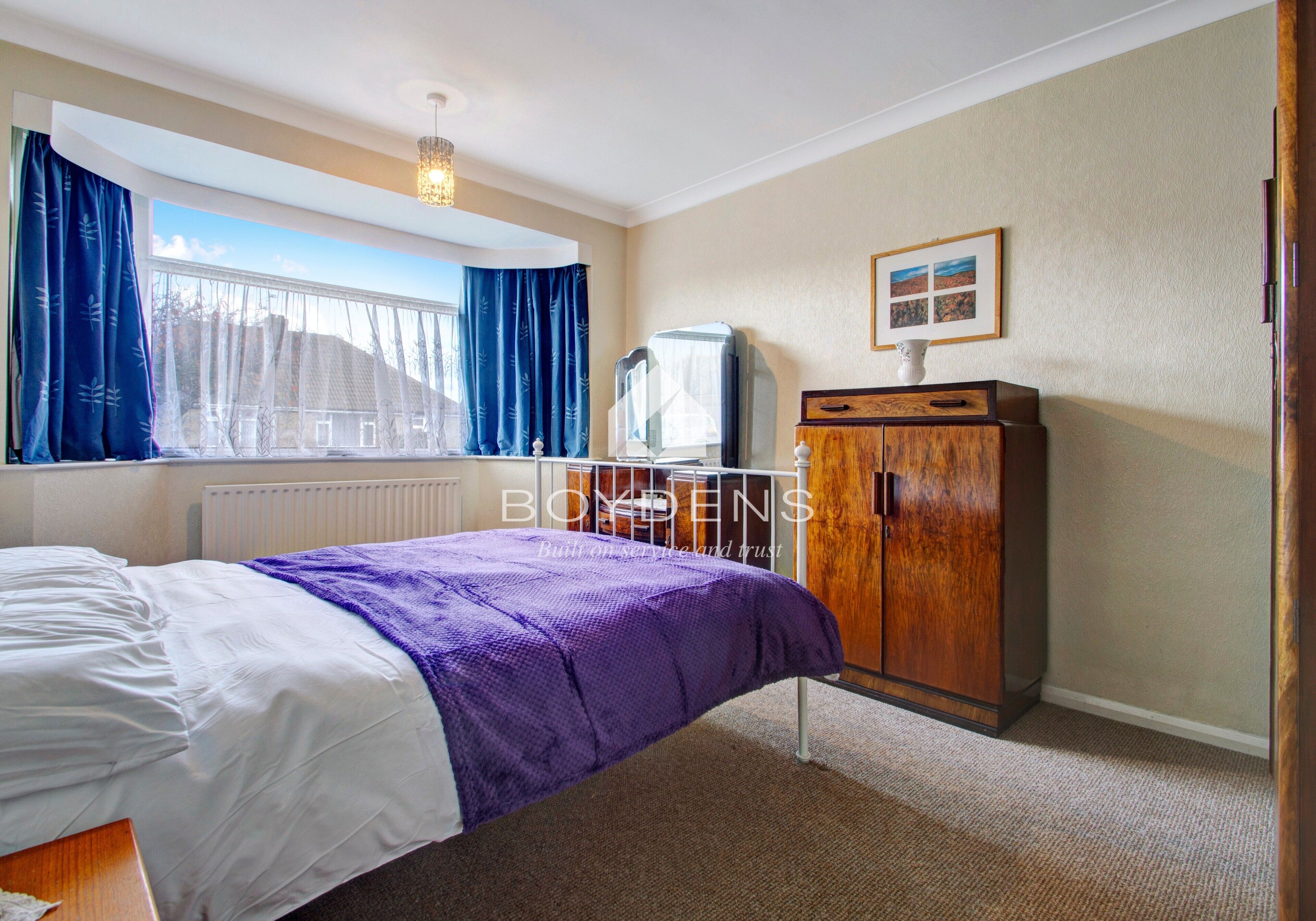 Une chambre à coucher avec un lit, une commode, un radiateur, une fenêtre avec des rideaux et un tableau accroché au mur.