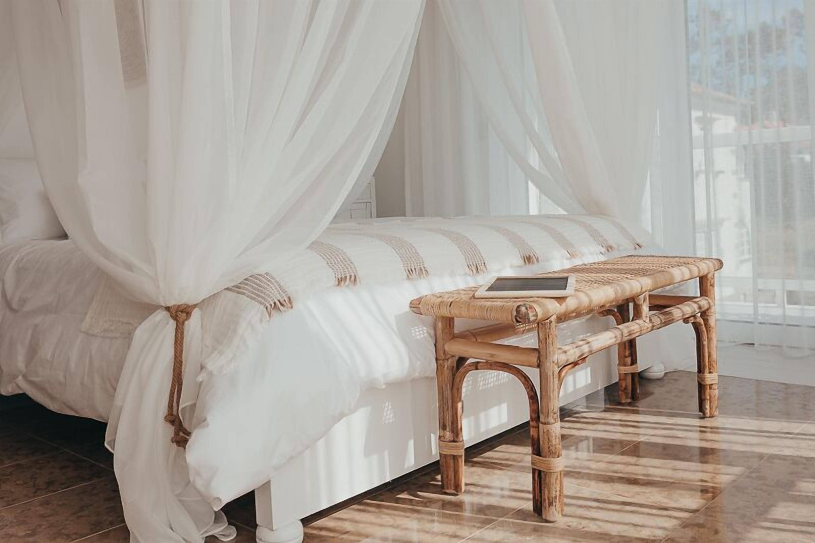A bedroom with a bed covered by white curtains, a wooden bench, and a bedside table.
