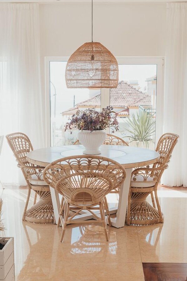 A round dining table with four wicker chairs, a hanging wicker pendant light, and a white vase with flowers. - Joy Beach House (Marinha Grande)