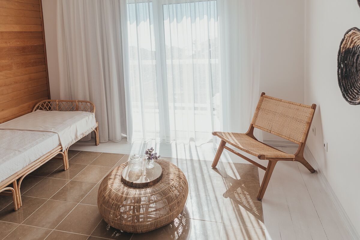 A room with a bed, a wicker chair, a round wicker coffee table, and a view through sheer curtains.