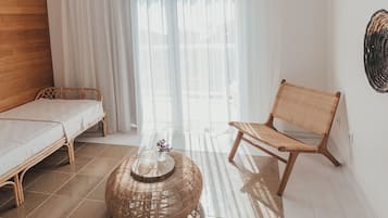 A room with a bed, a wicker chair, a round wicker coffee table, and a view through sheer curtains.