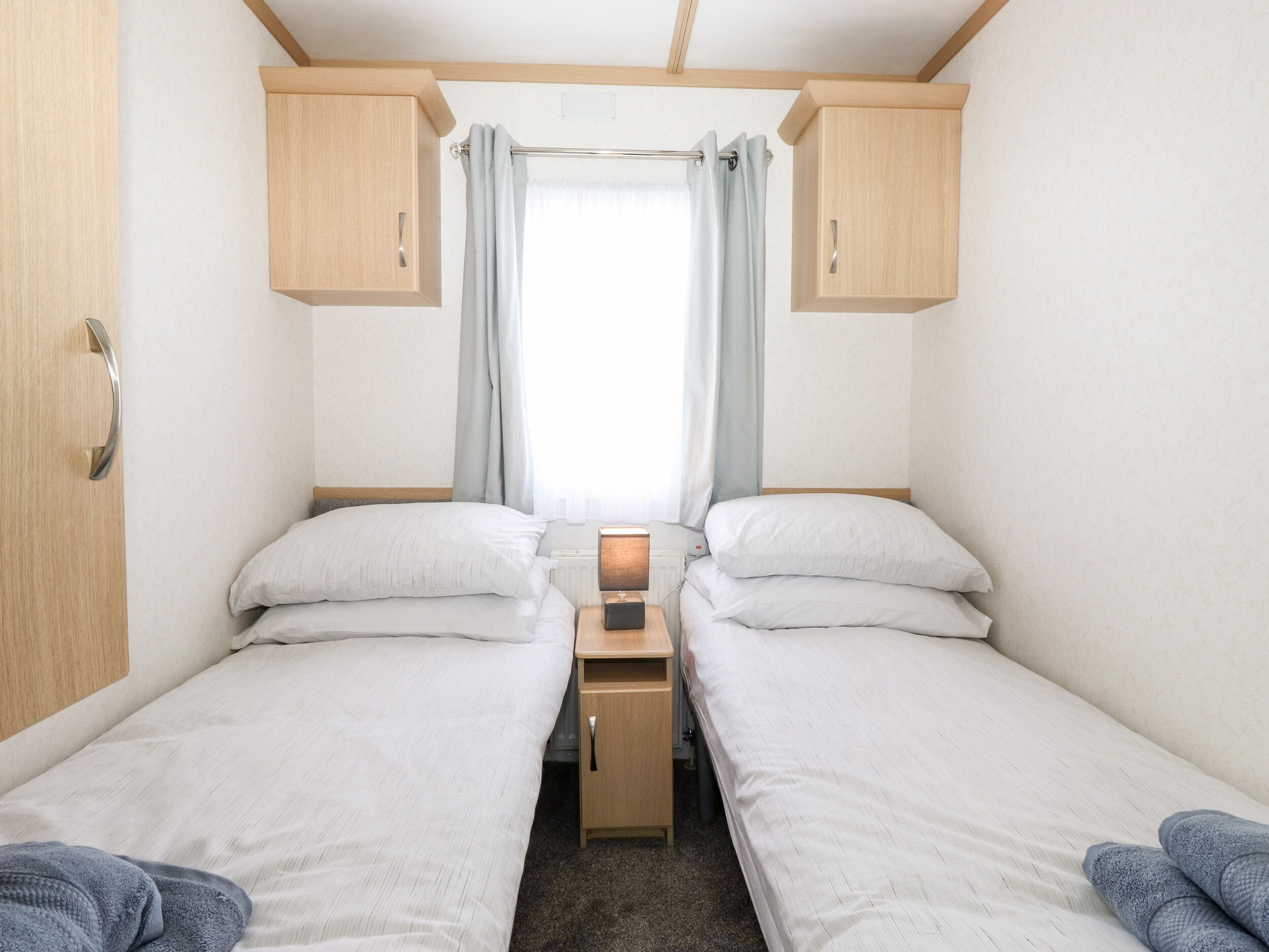 A compact bedroom with two single beds, wooden cabinets, a small table, and a window with curtains.