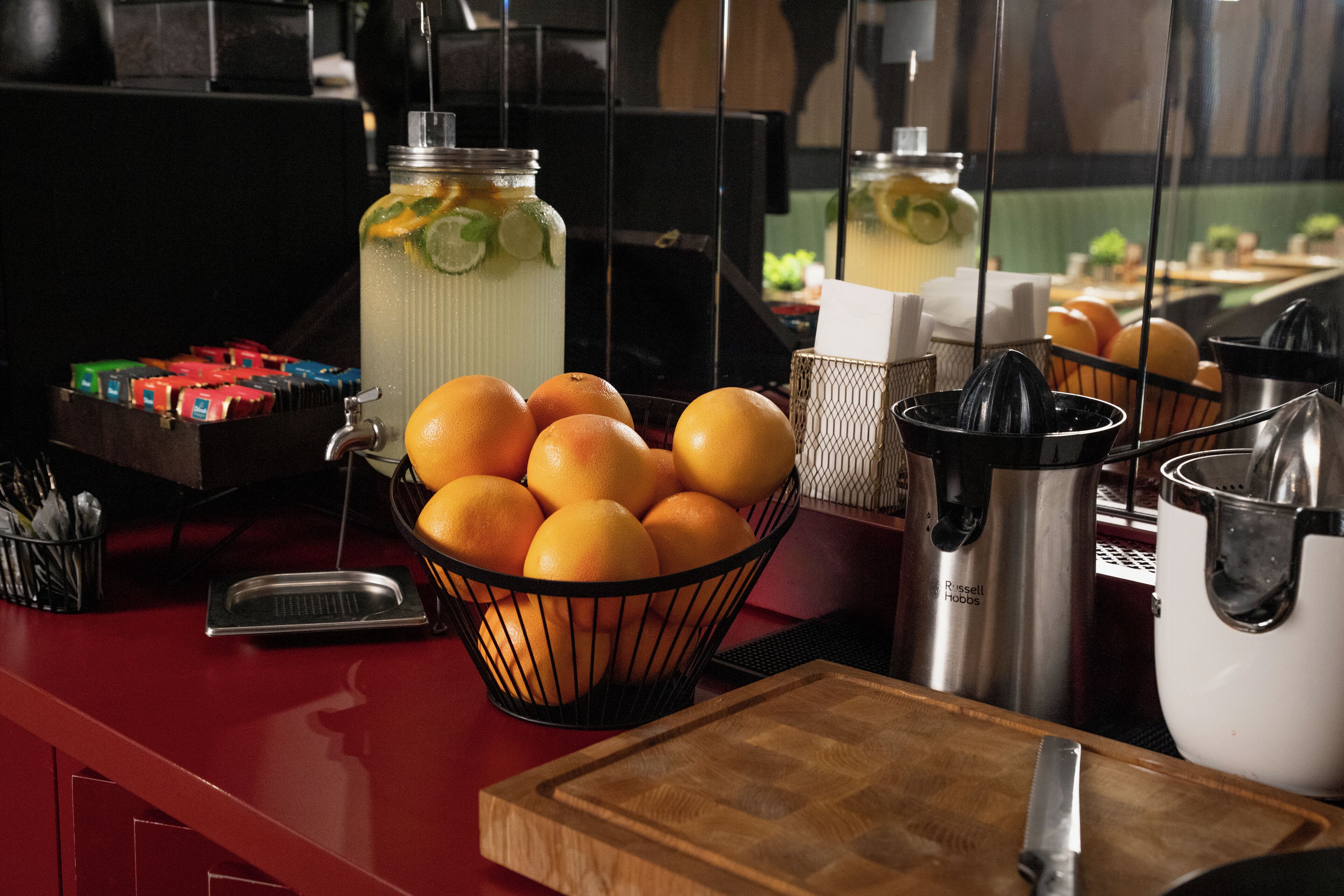 A kitchen counter with a basket of oranges, a juicer, a stainless steel pot, and a jar of lemonade.