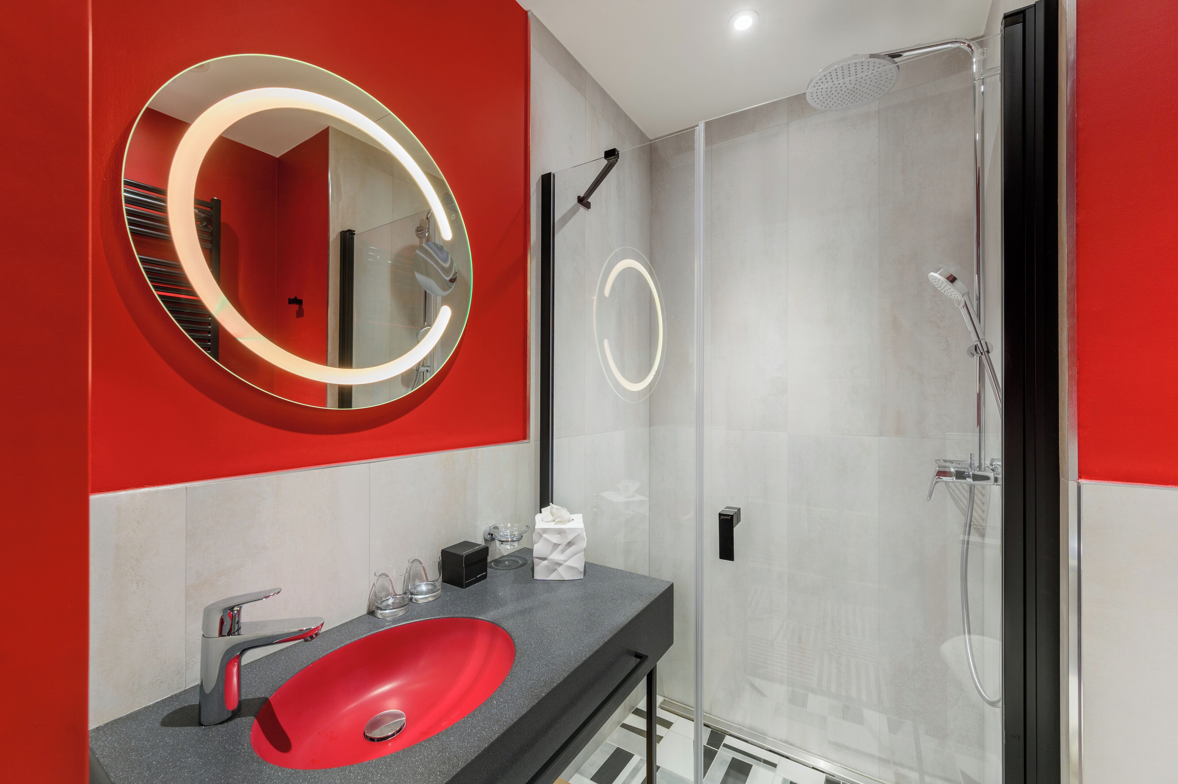 A modern bathroom with a red sink, a large mirror with a circular light, and a glass-enclosed shower.