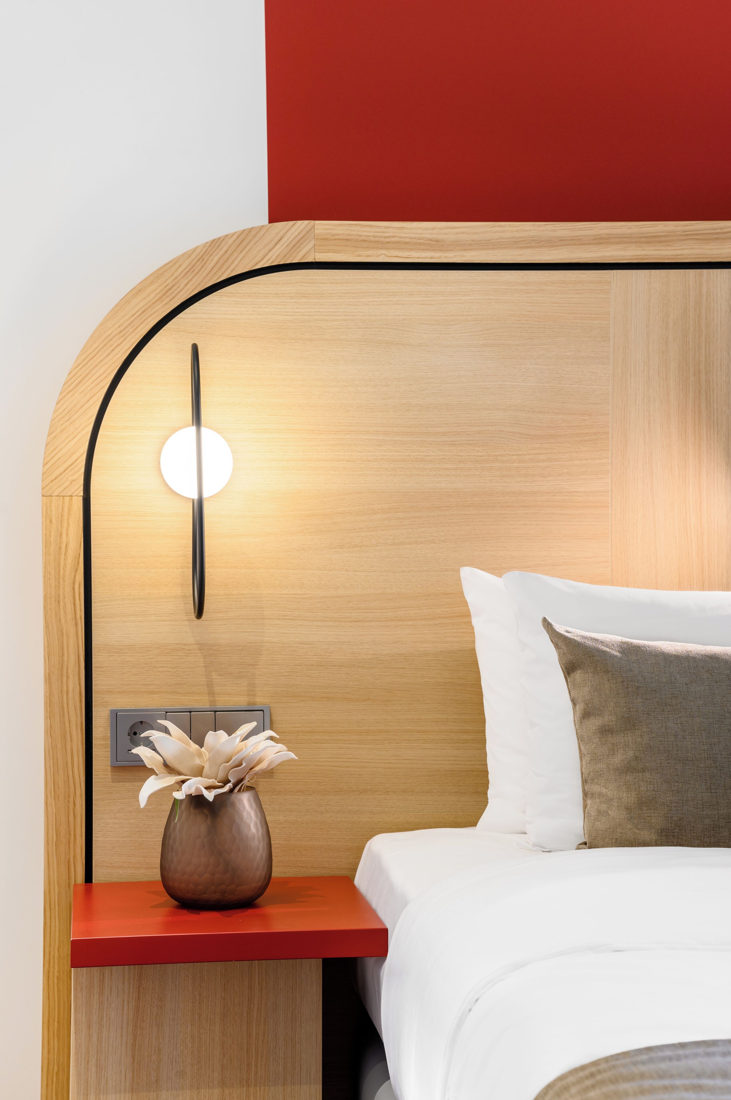 A modern hotel room with a wooden headboard, a bedside table with a vase of flowers, and a red accent wall.
