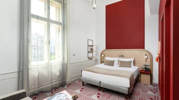 A hotel room with a large bed, a bedside table, a nightstand, a window with sheer curtains, and a red accent wall.