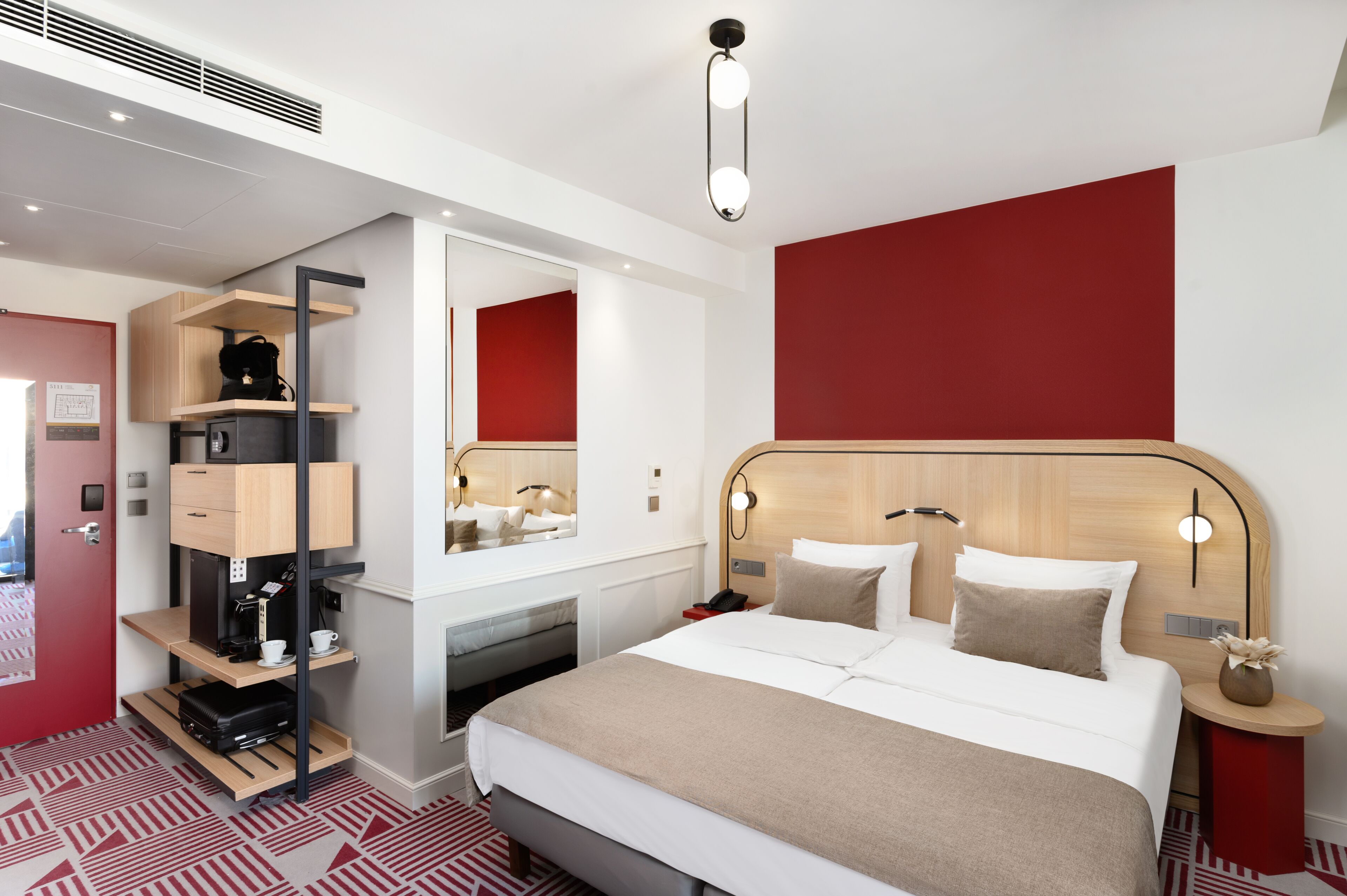 A modern hotel room with a large bed, a built-in desk with a mirror, and a red accent wall.
