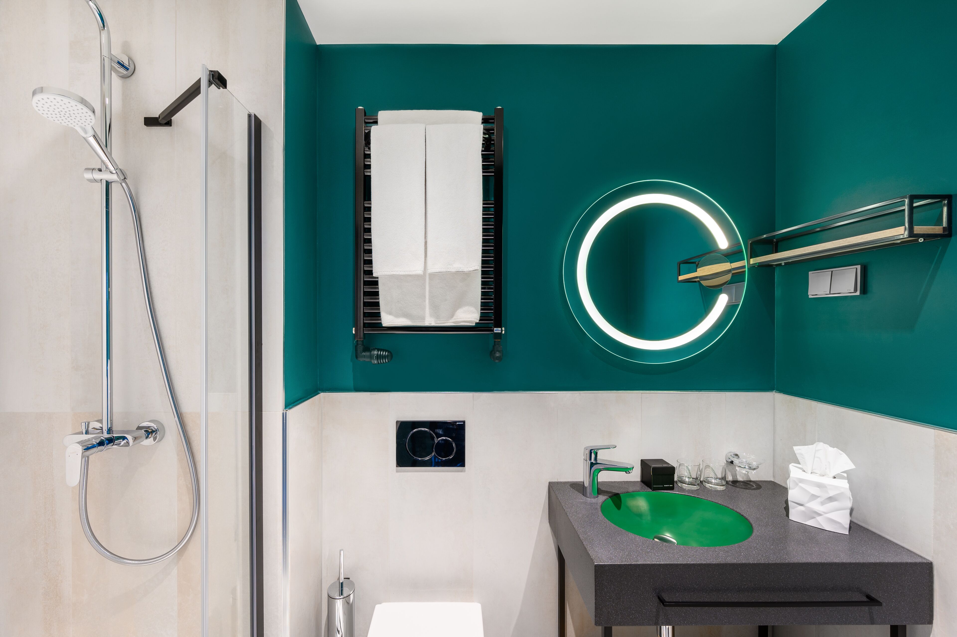A modern bathroom with a green sink, a towel rack, a mirror with a circular light, and a shower with a handheld showerhead.