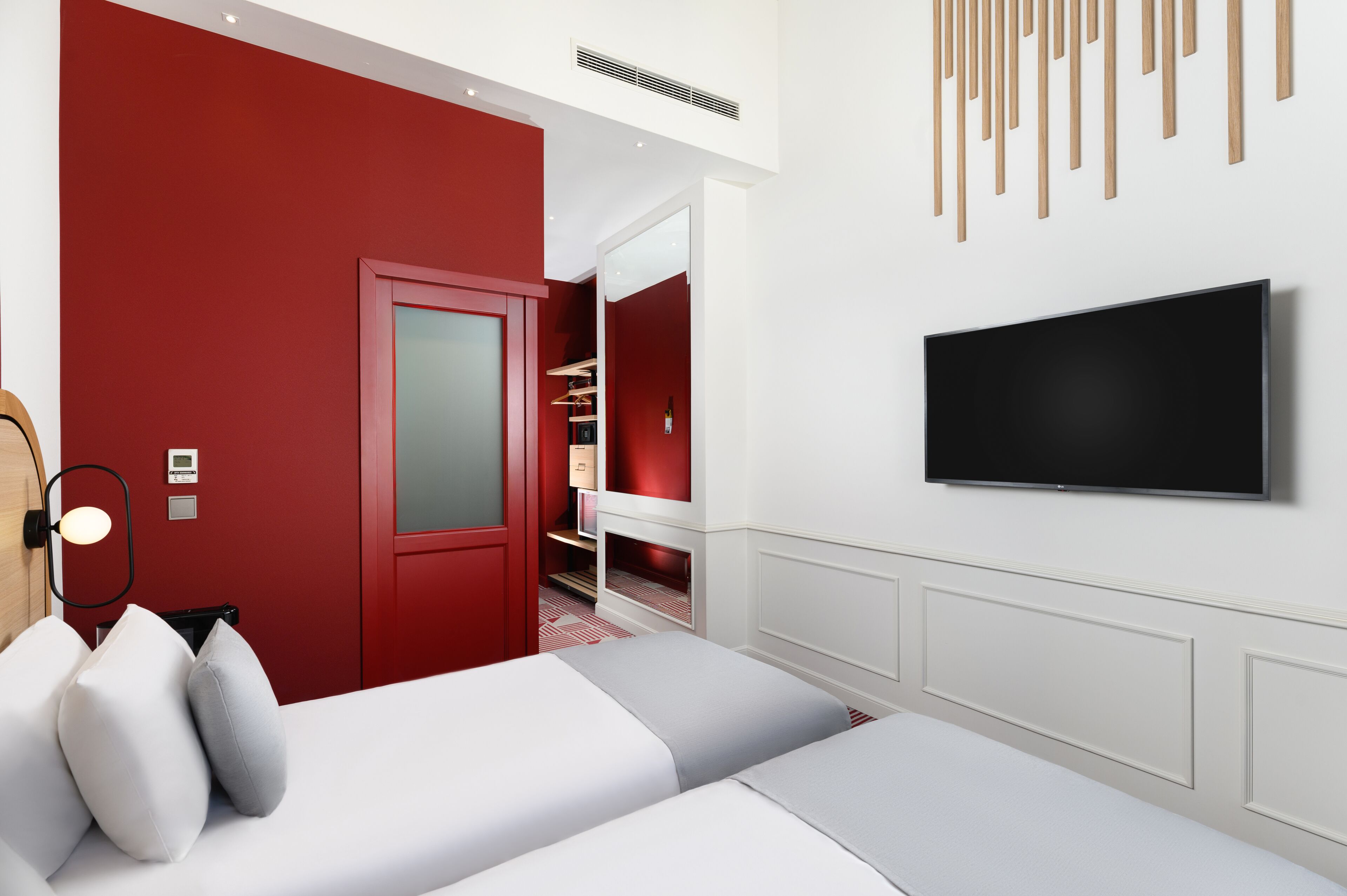 Una habitación de hotel moderna con una pared roja de acento, un televisor de pantalla plana, una cama con ropa de cama blanca y gris, y una mesita de noche con una lámpara.