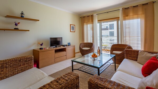 Ein modernes Wohnzimmer mit Glastisch, Korbmöbeln, einem Flachbildfernseher und einem Balkon mit Vorhängen.