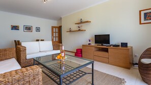 Un salon avec une table basse en verre, des meubles en osier, un canapé blanc, un meuble TV en bois et un téléviseur à écran plat.