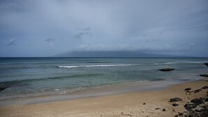 A beach with clear water, a sandy shore, and a mountain in the distance under a cloudy sky. - Pikake ocean front 20 feet from ocean (Lahaina)