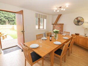 Un coin repas avec une table en bois dressée pour quatre personnes, entourée de chaises. Un mur de briques avec une fenêtre au-dessus de la table, et une horloge accrochée au mur, à droite.